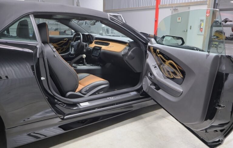 Gray sports car with open driver and passenger doors revealing tan leather interior and steering wheel in a service garage