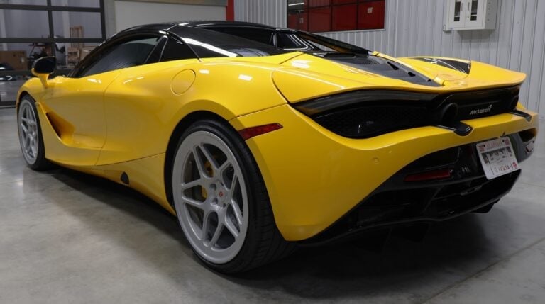 Bright yellow convertible sports car with black roof, white wheels, photographed from rear three-quarter view in industrial garage setting
